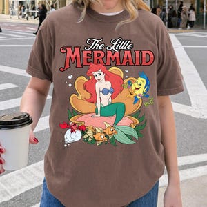May include: Brown t-shirt with a vibrant graphic of Ariel from The Little Mermaid. The design showcases Ariel, Flounder, Sebastian, and other sea creatures. The text "The Little Mermaid" is displayed above the graphic in a decorative font.