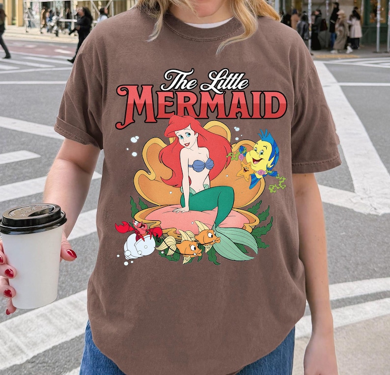 May include: Brown t-shirt with a vibrant graphic of Ariel from The Little Mermaid. The design showcases Ariel, Flounder, Sebastian, and other sea creatures. The text "The Little Mermaid" is displayed above the graphic in a decorative font.