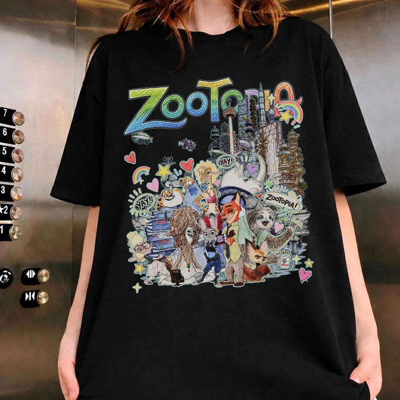 May include: Black t-shirt featuring a colorful graphic of characters and the cityscape from the animated film "Zootopia." The word "Zootopia" is written in a rainbow font above the illustration. The design includes various animals and buildings.
