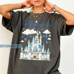 May include: Dark gray t-shirt featuring a graphic of Cinderella's castle with the text "Walt Disney World" in blue. The design includes clouds and blue orbs. The shirt is oversized.
