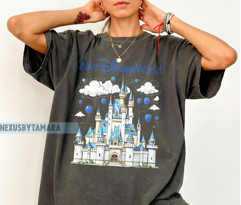 May include: Dark gray t-shirt featuring a graphic of Cinderella's castle with the text "Walt Disney World" in blue. The design includes clouds and blue orbs. The shirt is oversized.