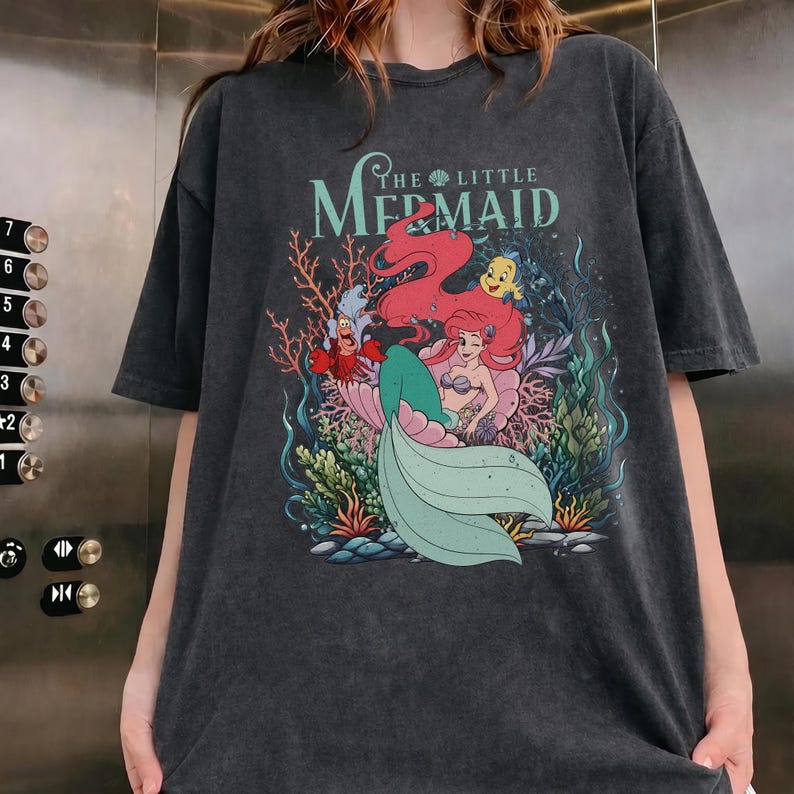 May include: Dark grey t-shirt with a colourful graphic from "The Little Mermaid." The design features Ariel with red hair, a green tail, and characters like Sebastian and Flounder. The text "The Little Mermaid" is above the characters.
