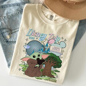 Puede incluir: Camiseta color crema con un dise&ntilde;o gr&aacute;fico colorido que presenta el texto "Disney Trip 2026" y una ilustraci&oacute;n de Baby Yoda sosteniendo una cabeza de Mickey Mouse. La camiseta tambi&eacute;n incluye im&aacute;genes de un castillo, una esfera y un &aacute;rbol.