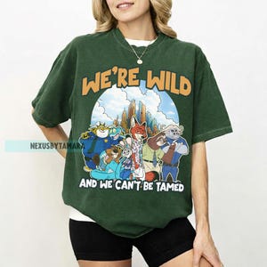 Puede incluir: Camiseta verde oscuro con el texto "WE'RE WILD" en naranja sobre una ilustraci&oacute;n de dibujos animados de personajes animales. Debajo, el texto "AND WE CAN'T BE TAMED" en blanco. La camiseta es de estilo casual.