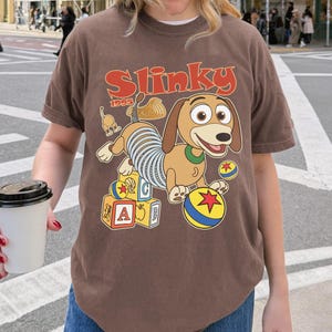 Puede incluir: Camiseta marr&oacute;n con una ilustraci&oacute;n de dibujos animados de Slinky Dog de Toy Story. El dise&ntilde;o incluye la palabra "Slinky" en naranja, el a&ntilde;o "1995" y bloques y bolas de colores. La camiseta la lleva una persona.