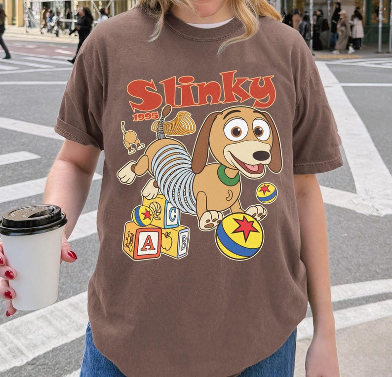 Puede incluir: Camiseta marr&oacute;n con una ilustraci&oacute;n de dibujos animados de Slinky Dog de Toy Story. El dise&ntilde;o incluye la palabra "Slinky" en naranja, el a&ntilde;o "1995" y bloques y bolas de colores. La camiseta la lleva una persona.