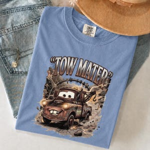 May include: Light blue t-shirt featuring a graphic of Tow Mater from the Cars movie. The design includes the text "TOW MATER" and an illustration of the character. The shirt is made of a soft material.
