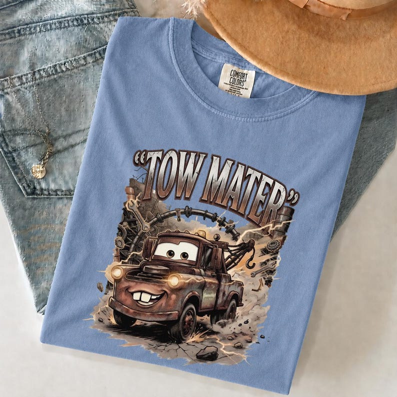 May include: Light blue t-shirt featuring a graphic of Tow Mater from the Cars movie. The design includes the text "TOW MATER" and an illustration of the character. The shirt is made of a soft material.