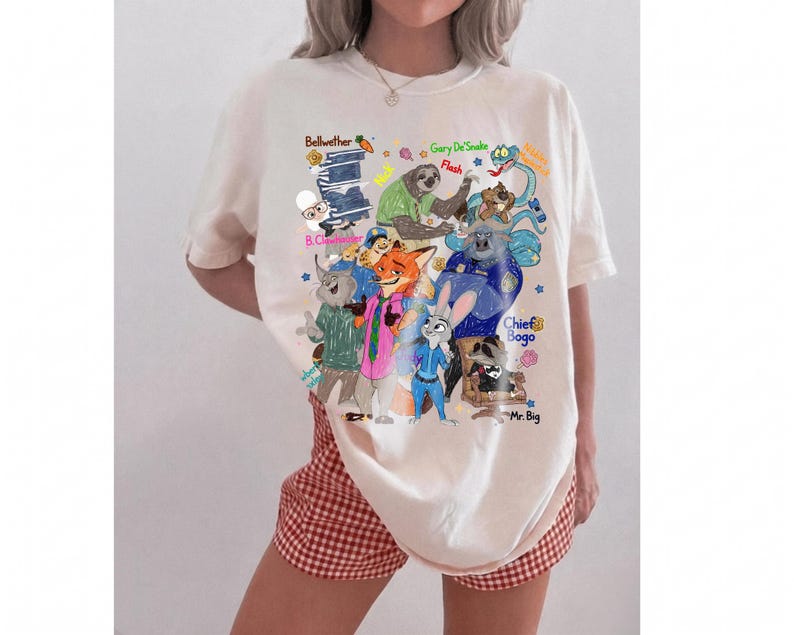 Op de afbeelding: Cr&egrave;mekleurig T-shirt met een kleurrijke afbeelding van verschillende stripfiguren met hun namen. Het ontwerp bevat personages als Bellwether, Gary DeSnake, Flash en Chief Bogo. Het shirt wordt door een persoon gedragen.