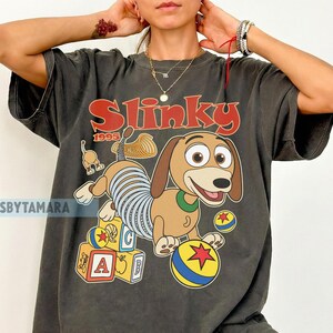 Puede incluir: Camiseta gris oscuro con un dise&ntilde;o de dibujos animados de Slinky Dog de Toy Story. El dise&ntilde;o incluye la palabra "Slinky" en rojo, el a&ntilde;o "1995" y bloques y bolas coloridas. La camiseta tiene un aspecto vintage y desgastado.