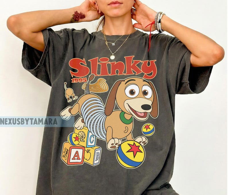 Puede incluir: Camiseta gris oscuro con un dise&ntilde;o de dibujos animados de Slinky Dog de Toy Story. El dise&ntilde;o incluye la palabra "Slinky" en rojo, el a&ntilde;o "1995" y bloques y bolas coloridas. La camiseta tiene un aspecto vintage y desgastado.