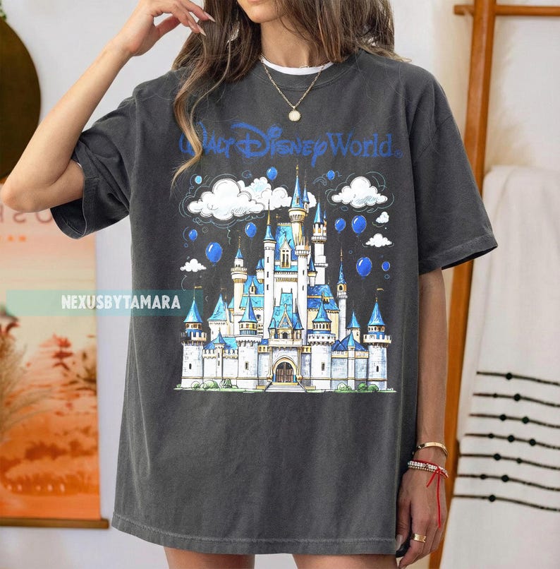 May include: Dark gray t-shirt featuring a blue and white illustration of a castle with the text "Walt Disney World". The design includes clouds and blue balloons. The shirt is a casual wear item.