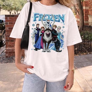 May include: White t-shirt with the word "FROZEN" in blue, and a graphic of characters from the animated film. The design includes Elsa, Anna, Kristoff, Olaf, and Sven. The shirt has a relaxed, oversized fit.