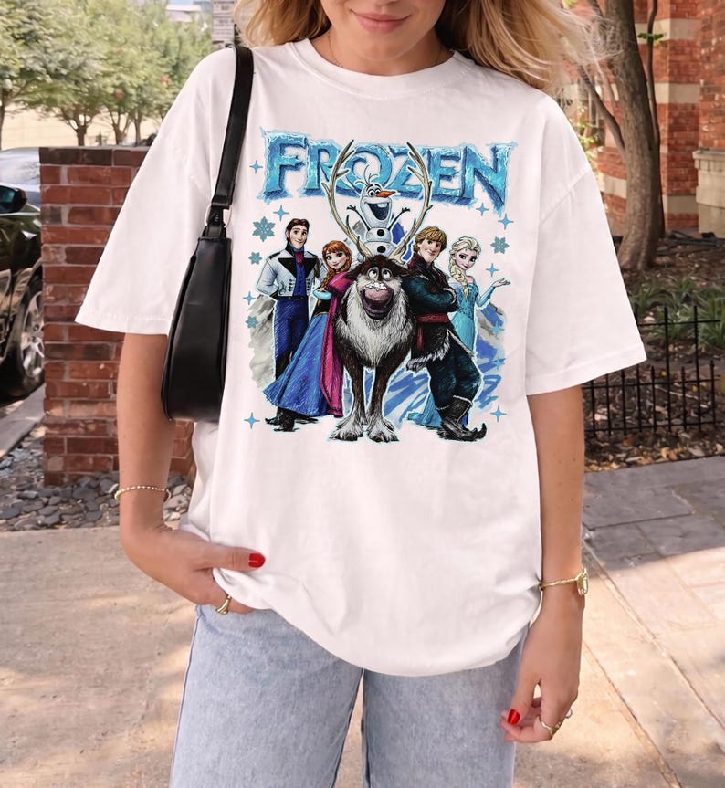 May include: White t-shirt with the word "FROZEN" in blue, and a graphic of characters from the animated film. The design includes Elsa, Anna, Kristoff, Olaf, and Sven. The shirt has a relaxed, oversized fit.