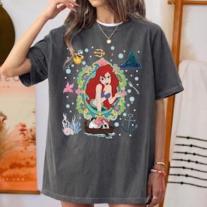 以下が含まれることがあります： 赤毛の人魚のカラフルなイラストが特徴の濃いグレーのTシャツ。水中の要素に囲まれています。デザインにはボート、城、錨が含まれています。ゆったりとしたフィット感のシャツです。