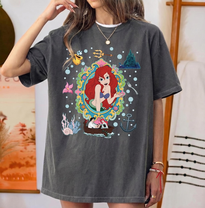 以下が含まれることがあります： 赤毛の人魚のカラフルなイラストが特徴の濃いグレーのTシャツ。水中の要素に囲まれています。デザインにはボート、城、錨が含まれています。ゆったりとしたフィット感のシャツです。