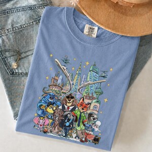 K&ouml;nnte beinhalten: Hellblaues Comfort Colors T-Shirt mit einer farbenfrohen Illustration einer Stadt mit Zeichentierfiguren. Das Design umfasst Wolkenkratzer, Hubschrauber und eine Einschienenbahn. Das T-Shirt liegt auf einer Jeans.