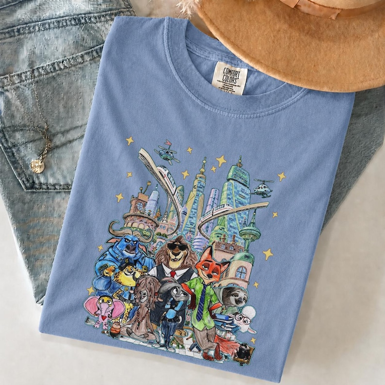 K&ouml;nnte beinhalten: Hellblaues Comfort Colors T-Shirt mit einer farbenfrohen Illustration einer Stadt mit Zeichentierfiguren. Das Design umfasst Wolkenkratzer, Hubschrauber und eine Einschienenbahn. Das T-Shirt liegt auf einer Jeans.