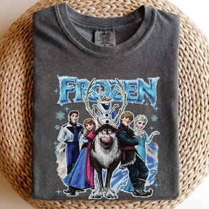 May include: Dark grey t-shirt with the word "FROZEN" in blue, and images of characters from the animated film. The design includes Elsa, Anna, Kristoff, Olaf, and Sven. The shirt is made of Comfort Colors material.