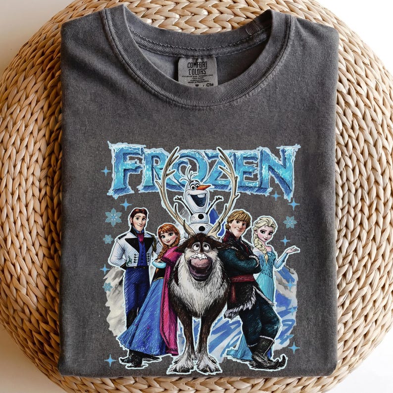 May include: Dark grey t-shirt with the word "FROZEN" in blue, and images of characters from the animated film. The design includes Elsa, Anna, Kristoff, Olaf, and Sven. The shirt is made of Comfort Colors material.