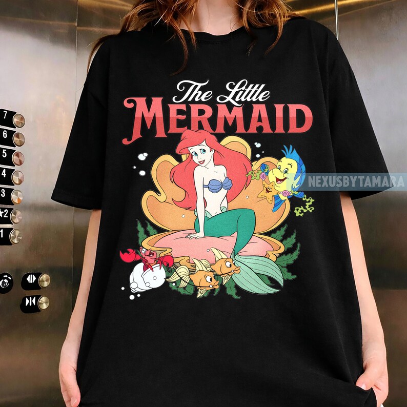 May include: Black t-shirt with a colourful graphic of Ariel from The Little Mermaid. The design includes Ariel, Flounder, Sebastian, and other sea creatures. The words "The Little Mermaid" are printed above the graphic.