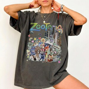 May include: Dark gray t-shirt featuring the word "Zootopia" in colorful lettering. The shirt showcases a vibrant graphic of characters and cityscapes from the animated film, with additional text and illustrations.