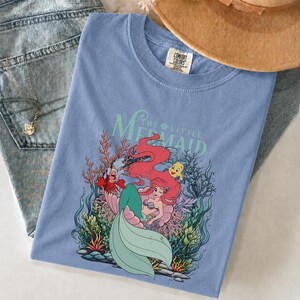 May include: A pale blue t-shirt with a graphic of Ariel from The Little Mermaid. The design features Ariel with red hair, a green tail, and other characters. The words "The Little Mermaid" are printed above the image.