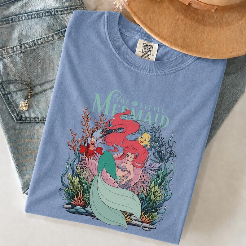 May include: A pale blue t-shirt with a graphic of Ariel from The Little Mermaid. The design features Ariel with red hair, a green tail, and other characters. The words "The Little Mermaid" are printed above the image.