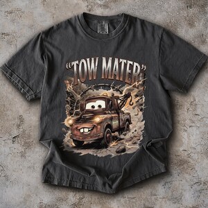 May include: Dark gray t-shirt featuring a cartoon image of Tow Mater from the movie Cars. The graphic shows Mater with a smiling face, headlights, and the text "TOW MATER". The design includes lightning bolts and mechanical elements.