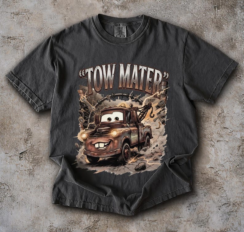 May include: Dark gray t-shirt featuring a cartoon image of Tow Mater from the movie Cars. The graphic shows Mater with a smiling face, headlights, and the text "TOW MATER". The design includes lightning bolts and mechanical elements.