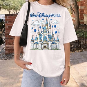 May include: White t-shirt featuring a blue and white illustration of a castle with the text "Walt Disney World." The design includes blue balloons and clouds. The shirt has short sleeves and a relaxed fit.
