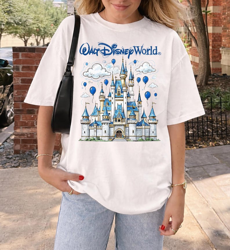 May include: White t-shirt featuring a blue and white illustration of a castle with the text "Walt Disney World." The design includes blue balloons and clouds. The shirt has short sleeves and a relaxed fit.
