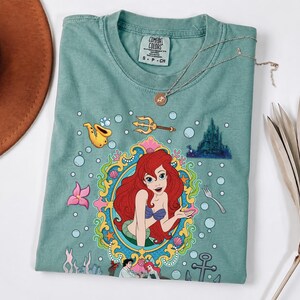 以下が含まれることがあります： 人魚姫のアリエルのカラフルなイラストが描かれた、シーフォームグリーンのTシャツ。デザインにはアリエルの肖像画、お城、その他の水生要素が含まれています。金のネックレスがシャツの上にかけられています。