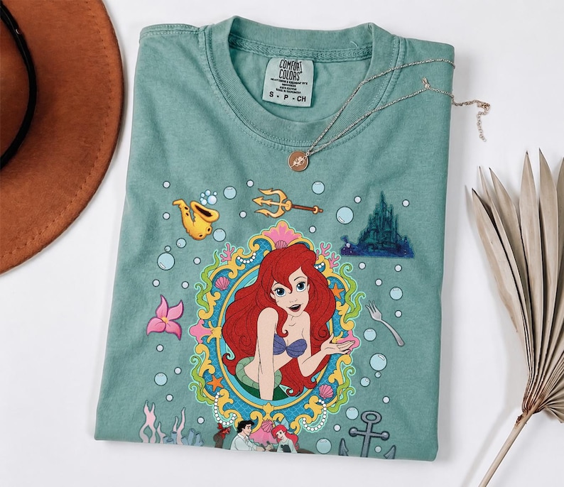 以下が含まれることがあります： 人魚姫のアリエルのカラフルなイラストが描かれた、シーフォームグリーンのTシャツ。デザインにはアリエルの肖像画、お城、その他の水生要素が含まれています。金のネックレスがシャツの上にかけられています。
