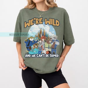 Puede incluir: Camiseta verde oliva con un gr&aacute;fico de dibujos animados de personajes de animales y el texto "WE'RE WILD AND WE CAN'T BE TAMED". El dise&ntilde;o incluye un tel&oacute;n de fondo de paisaje urbano. La camiseta es de estilo casual y de gran tama&ntilde;o.