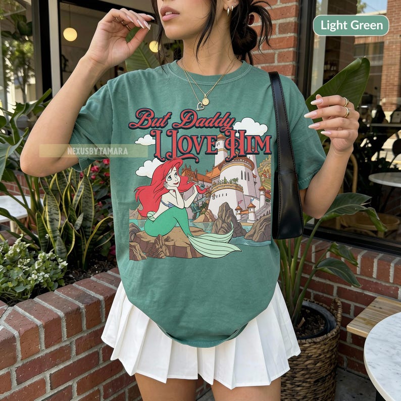 Puede incluir: Camiseta verde claro con un gr&aacute;fico de Ariel de La Sirenita, con el texto "But Daddy I Love Him". El dise&ntilde;o incluye un castillo y nubes. La camiseta es de color verde claro.