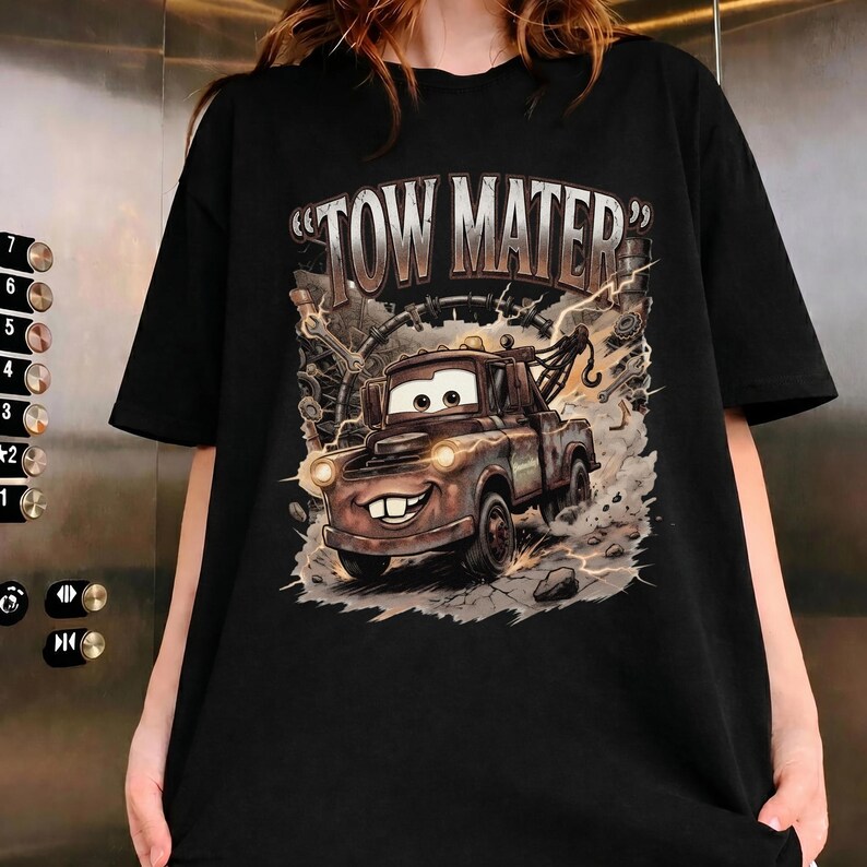May include: Black t-shirt featuring a cartoon image of Tow Mater from the movie Cars. The graphic shows Mater with a big smile, surrounded by mechanical elements and the text "TOW MATER" in a distressed font.