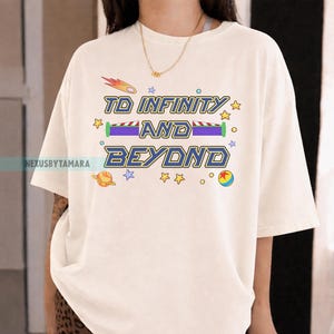 May include: Cream-colored t-shirt with the phrase "TO INFINITY AND BEYOND" in blue and yellow lettering. The design includes a shooting star, planets, stars, and a toy ball. The shirt is a relaxed fit.