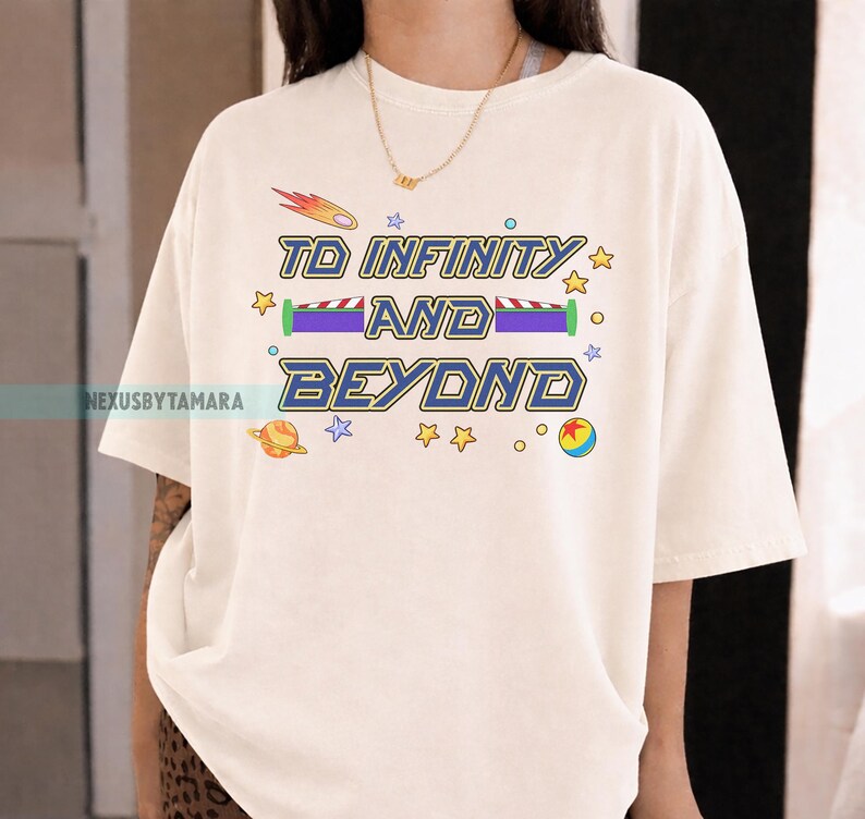 May include: Cream-colored t-shirt with the phrase "TO INFINITY AND BEYOND" in blue and yellow lettering. The design includes a shooting star, planets, stars, and a toy ball. The shirt is a relaxed fit.