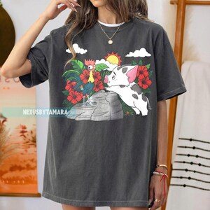 May include: Dark gray t-shirt featuring a colorful graphic of a rooster and a pig, set against a tropical backdrop with red flowers, a sun, and clouds. The text "NexusbyTamara" is printed on the shirt.