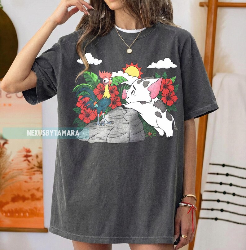 May include: Dark gray t-shirt featuring a colorful graphic of a rooster and a pig, set against a tropical backdrop with red flowers, a sun, and clouds. The text "NexusbyTamara" is printed on the shirt.