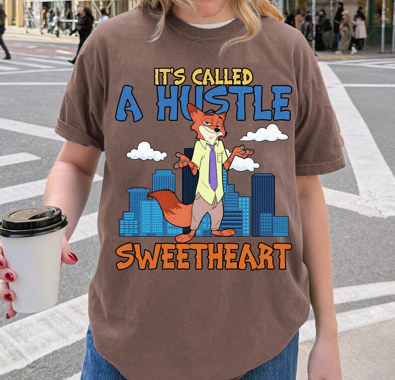 以下が含まれることがあります： 「IT'S CALLED A HUSTLE SWEETHEART」の文字と、シャツとネクタイを着た漫画のキツネのキャラクターが描かれた茶色のTシャツ。キツネは青い建物と白い雲を背景に立っています。