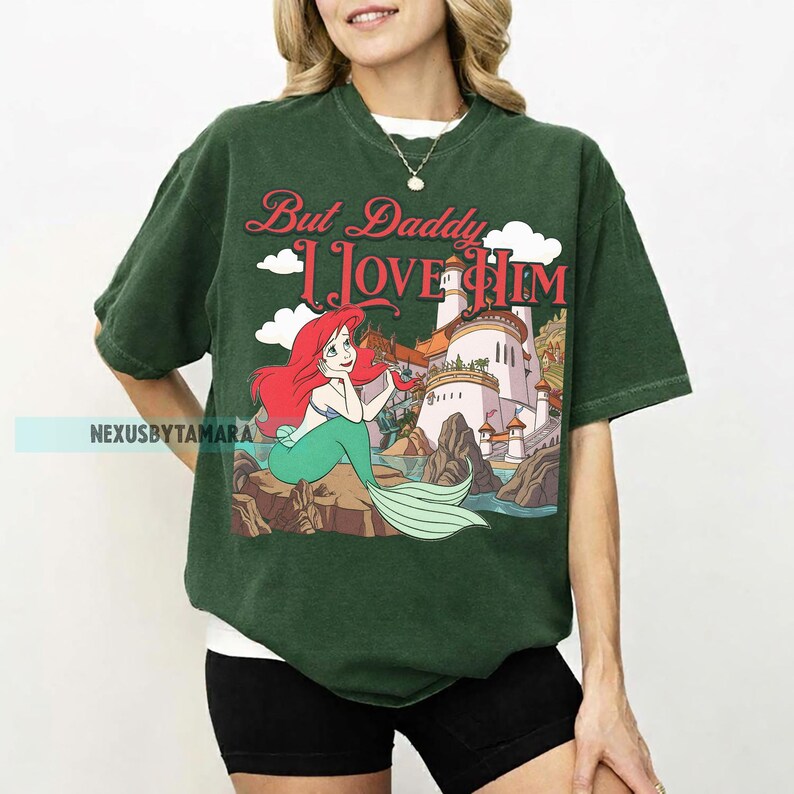 Puede incluir: Camiseta verde oscuro con un gr&aacute;fico de Ariel de La Sirenita, con el texto "But Daddy I Love Him". La ilustraci&oacute;n muestra a Ariel en una roca con un castillo al fondo. La camiseta es de corte holgado.