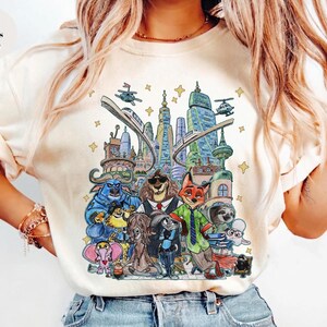 K&ouml;nnte beinhalten: Elfenbeinfarbenes T-Shirt mit einer farbenfrohen Illustration von Zootopia-Charakteren und Stadtlandschaften. Das Design zeigt verschiedene Tierfiguren, Geb&auml;ude und Verkehrselemente. Der Text "Colors Ivory" befindet sich oben links.