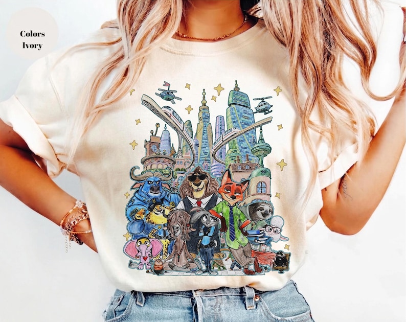K&ouml;nnte beinhalten: Elfenbeinfarbenes T-Shirt mit einer farbenfrohen Illustration von Zootopia-Charakteren und Stadtlandschaften. Das Design zeigt verschiedene Tierfiguren, Geb&auml;ude und Verkehrselemente. Der Text "Colors Ivory" befindet sich oben links.