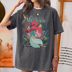 May include: Dark grey t-shirt with a graphic of Ariel from The Little Mermaid. The design features Ariel with red hair, a green tail, and other characters. The text "The Little Mermaid" is arched above the image.