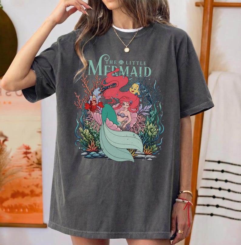 May include: Dark grey t-shirt with a graphic of Ariel from The Little Mermaid. The design features Ariel with red hair, a green tail, and other characters. The text "The Little Mermaid" is arched above the image.