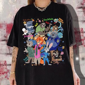 Op de afbeelding: Zwart T-shirt met een kleurrijke illustratie van personages uit de animatiefilm Zootopia. Het ontwerp bevat verschillende dierenpersonages met hun namen, zoals Judy, Nick en Flash. Het shirt is een casual kledingstuk.