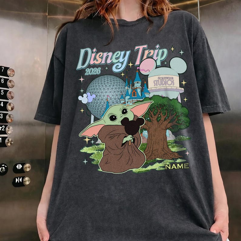 Puede incluir: Camiseta gris oscuro con un gr&aacute;fico de Baby Yoda sosteniendo un peluche de Mickey Mouse, con el texto "Disney Trip 2026". El dise&ntilde;o incluye una escena de parque con un &aacute;rbol, un castillo y una torre de agua.