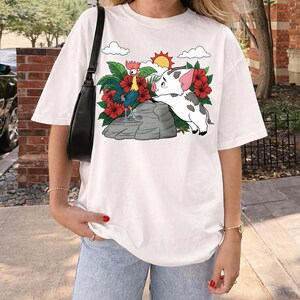 May include: White t-shirt featuring a colorful graphic of a rooster and a pig, surrounded by red flowers, green leaves, clouds, and a sun. The design is set against a light gray rock. The shirt is a casual, oversized style.